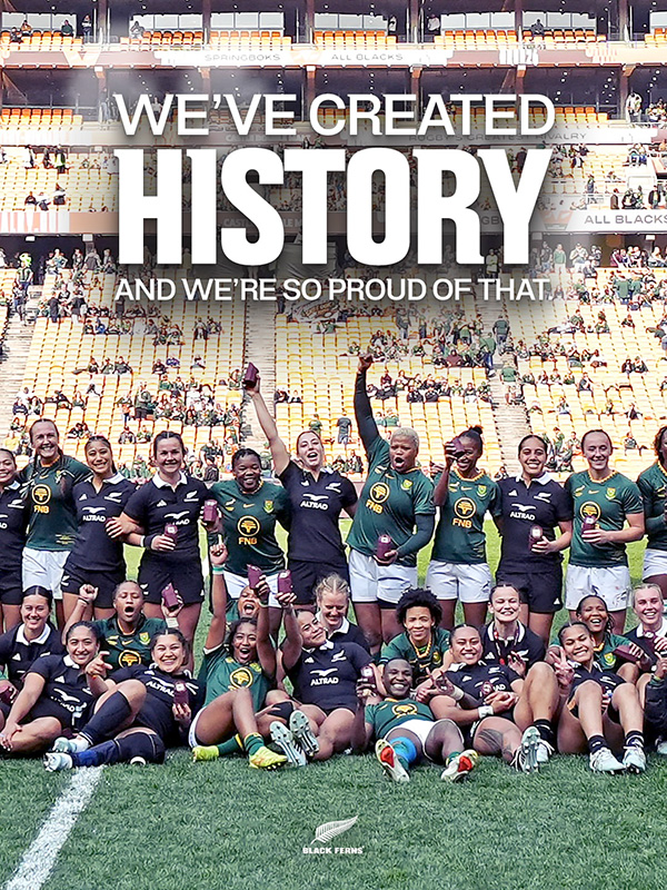 Springbok Women and Black Ferns players pose together on the FNB Stadium pitch after their historic first Test on South African soil, beneath the words We've created history and we're so proud of that. Players in green and gold and in black stand and sit side by side, smiling and celebrating, several holding small maroon commemorative boxes, with the Black Ferns logo at the foot of the graphic.